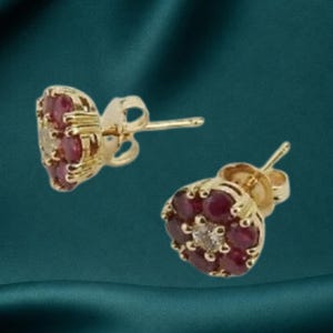 May include: A pair of gold stud earrings featuring a floral cluster design. Each earring consists of a central clear stone surrounded by six round red stones, all set in a gold metal frame with a standard post and butterfly back closure mechanism.