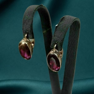 May include: A pair of drop earrings featuring oval-shaped, deep magenta-coloured gemstones set in polished gold-toned metal frames. The earrings are suspended from curved, dark grey display stands, showcasing the faceted cut of the stones.