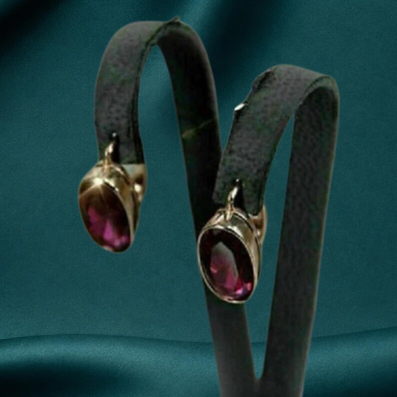 May include: A pair of drop earrings featuring oval-shaped, deep magenta-coloured gemstones set in polished gold-toned metal frames. The earrings are suspended from curved, dark grey display stands, showcasing the faceted cut of the stones.