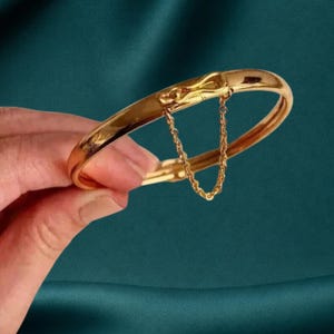 May include: A polished gold-tone hinged bangle bracelet featuring a smooth, rounded surface and a delicate, thin gold chain connecting the two ends of the clasp mechanism. The bracelet has a secure locking closure and a high-shine metallic finish.
