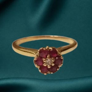 May include: A gold ring featuring a floral-shaped cluster setting. The center of the flower is a single clear stone surrounded by six round, deep red rubies. The band is a smooth, polished gold metal that tapers slightly toward the central setting.