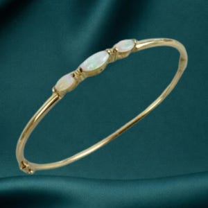 May include: A thin gold bangle bracelet featuring three marquise-cut opals set in a row along the top. The metal band is smooth and polished, creating a continuous circular shape. The opals display iridescent flashes of color against the gold setting.