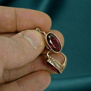 May include: A gold-toned metal pendant featuring two oval-shaped, deep red faceted gemstones. The upper stone is set within a smooth metal frame, while the lower stone is held in a matching metal setting, creating a linked, vertical drop design.