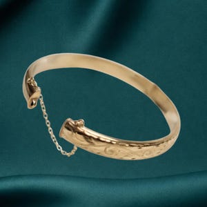 Könnte beinhalten: Ein goldfarbener Klapparmreif mit eingraviertem Blumenmuster auf der Außenseite. Das Armband verfügt über eine zarte Sicherheitskette, die die beiden Enden verbindet, welche durch einen Verschlussmechanismus an einer Seite gesichert sind.