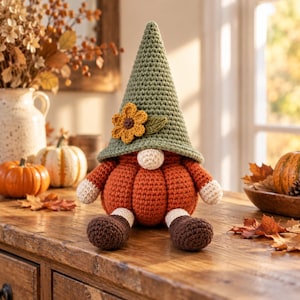May include: A crocheted gnome figure featuring a tall, sage green pointed hat adorned with a yellow flower and a green leaf. The body is shaped like a round, orange pumpkin with cream-colored hands and dark brown feet, all crafted in yarn.
