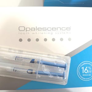 May include: A white and blue box for Opalescence tooth whitening systems. The box features the brand name, a row of six gray circles, and a circular seal stating 16% PF Regular with potassium nitrate and fluoride. Two syringes are visible.