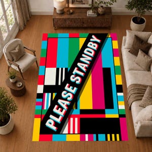 Op de afbeelding: Een rechthoekig vloerkleed met een levendig televisie-testbeeldontwerp met verticale en horizontale blokken in rood, geel, blauw, groen, zwart en wit. Een brede zwarte diagonale streep toont de witte tekst PLEASE STANDBY.