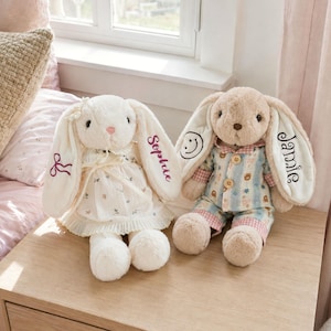 May include: Two plush rabbit toys with long ears. The left rabbit is cream-colored, wearing a floral dress with a bow, and has the name Sophie embroidered on its ear. The right rabbit is tan, wearing blue striped pajamas, with a smiley face and the name Jamie embroidered on its ears.