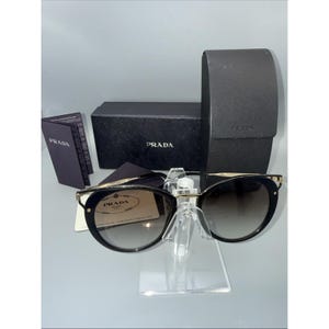 May include: Black Prada sunglasses with gold-tone metal accents and gradient lenses. The set includes a black textured Prada hard case, a black Prada box, a small Prada information booklet, and a Prada authenticity card with the brand logo.