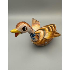 Pode incluir: Um brinquedo de lata em forma de pato com corpo às riscas castanhas e bege, bico amarelo e fita azul no pescoço. O pato tem asas estendidas com padrões de penas detalhados e olhos grandes e expressivos, feito de componentes de metal pintado.