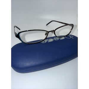 May include: Dark brown eyeglasses with thin frames and a subtle teal accent on the outer edges, resting on a smooth, oval-shaped blue hard case. The case features a white floral graphic and the letters OP printed on the top surface.