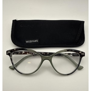 May include: Cat-eye glasses with a gray and black tortoiseshell pattern on the frames and clear lenses. The glasses are positioned in front of a black fabric protective case with a white tag that reads MODFANS in capital letters.