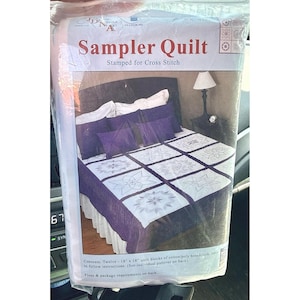 May include: A Sampler Quilt stamped for cross stitch kit in plastic packaging. The package features an image of a bed with a white and purple quilt, purple pillows, and a lamp. Text on the package reads Sampler Quilt Stamped for Cross Stitch.