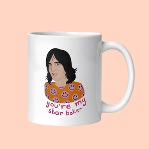 May include: A white ceramic mug featuring a graphic illustration of a person with dark hair wearing an orange shirt with a pink smiley face pattern. Below the illustration, the text reads you're my star baker in pink lowercase letters.