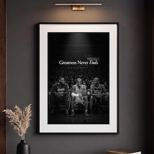 May include: A framed black and white wall art print featuring three basketball players sitting on a bench in a stadium. The text Greatness Never Ends is printed in white in the center. The print is displayed in a thin black frame on a dark wall.