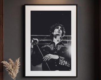 May include: A black and white framed photograph of a person with curly hair wearing a button-down shirt with intricate embroidery on the chest and cuffs, holding a microphone stand while looking upward in a performance setting.