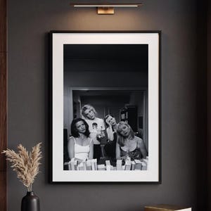 Op de afbeelding: Een zwart-wit ingelijste wandkunstprint met een groep van vier personen in een kamer. De print wordt weergegeven in een dunne zwarte lijst met een brede witte passe-partout. De afbeelding toont de onderwerpen in een informele setting met diverse items.