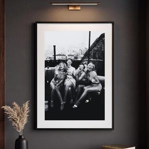Pu&ograve; includere: Una stampa artistica da parete incorniciata in bianco e nero che raffigura quattro persone che ridono insieme su una terrazza sul tetto. La stampa rettangolare &egrave; inserita in una sottile cornice nera ed &egrave; montata su una parete grigio scuro sopra un mobile in legno.