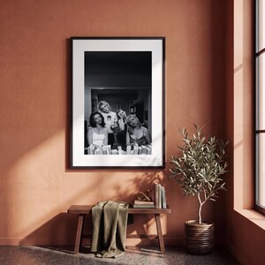 Op de afbeelding: Een zwart-wit ingelijste foto met vier personen in een kamer, gepresenteerd in een dunne zwarte lijst met een witte passe-partout. De afbeelding toont een groep mensen in een informele setting, gecentreerd binnen de lijst.