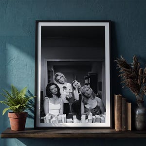 Op de afbeelding: Een zwart-wit ingelijste wandkunstprint met een groep van vier personen in een kamer. De afbeelding toont drie zittende personen en &eacute;&eacute;n staande persoon, met een rij boeken op de voorgrond. De lijst is dun, zwart en heeft een witte passe-partout.