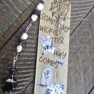 Puede incluir: Un marcapáginas de madera con una telaraña pirograbada y el texto "Something wicked this way comes". Está decorado con pegatinas de una botella, una polilla y una calavera, e incluye una borla con cuentas de calavera blancas y gemas moradas.