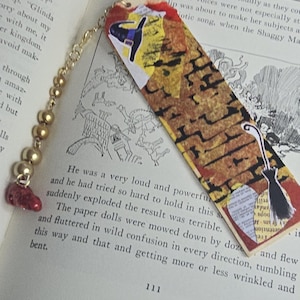 May include: A rectangular bookmark featuring a collage of yellow, orange, and brown tones with a witch hat and broomstick illustration. A gold-toned chain with metallic beads and a small red charm is attached to the top of the bookmark.