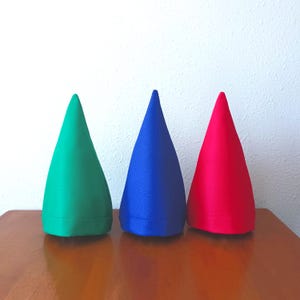 May include: Three cone-shaped fabric ornaments in solid green, blue, and red colors. Each piece features a smooth, matte texture with a visible stitched hem along the bottom edge, standing upright in a row against a plain white background.