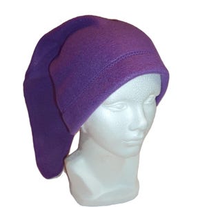 May include: A solid purple fleece hat with a long, pointed, drooping tail that hangs down the side. The hat features a wide, folded brim with visible stitching along the edge, constructed from a soft, matte fabric material.
