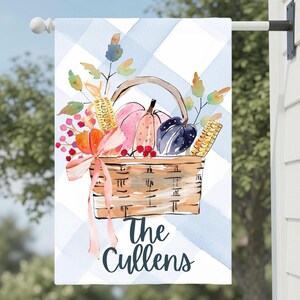 May include: A decorative garden flag featuring a watercolor illustration of a woven basket filled with pumpkins, corn, and berries. A light pink ribbon is tied to the handle. The background is a light blue and white plaid pattern with the text The Cullens.
