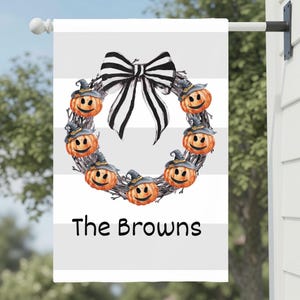 May include: A rectangular garden flag featuring a horizontal white and light gray striped background. A wreath made of twigs is decorated with six orange pumpkins wearing black witch hats and a large black and white striped bow. The text The Browns is printed in black below the wreath.