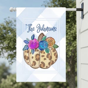 Könnte beinhalten: Eine rechteckige Gartenflagge mit einem hellblau-weißen Karomuster. In der Mitte befindet sich ein Kürbis mit Leopardenmuster, der mit bunten rosa, blauen und orangefarbenen Blumen verziert ist. Der Text The Johnsons ist in Blau gedruckt.