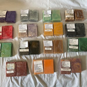 May include: A collection of rectangular soap bars in various colors including purple, red, green, brown, orange, and black. Each bar is individually wrapped in clear plastic and features a white paper label with text from Amy's Afghans and Things.