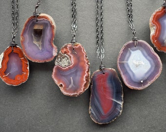May include: Seven necklaces featuring polished agate slices in shades of orange, red, purple, and white. Each stone is suspended from a dark metal chain by a wire-wrapped bail, showcasing natural banding and crystalline centers.