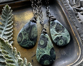 May include: Three teardrop-shaped pendants made of green and black patterned stone, each suspended from a dark metal chain. The stones feature circular, eye-like markings and are secured at the top with wrapped dark metal wire.