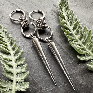 May include: A pair of silver-colored, long, pointed earrings. Each earring features a hoop, a woven metal design, another hoop, and a long, conical spike. The earrings are displayed on a dark gray surface with green leafy accents.