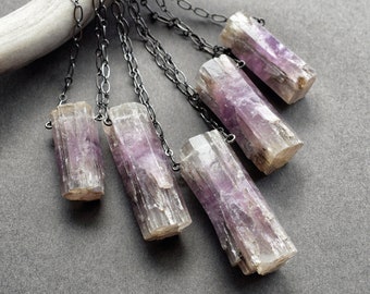 May include: Six necklaces featuring rectangular, pale purple and clear crystal pendants. The pendants hang from dark grey metal chains. The crystals have a rough, natural texture. The background is a solid grey.