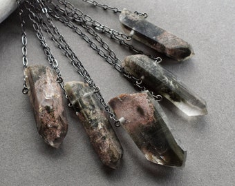 May include: A collection of crystal pendant necklaces. Each necklace has a clear, elongated crystal with dark green and pink inclusions, hanging from a dark silver chain. The crystals vary in size and colour, set against a grey backdrop.