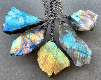 May include: Five raw labradorite pendants suspended on dark metal chains. Each stone features a unique, irregular shape with iridescent flashes of blue, gold, and green. The stones are secured with dark wire wrapping at the top.