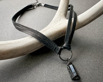 Könnte beinhalten: Eine schwarze Lederkette mit einem silbernen Ring und einem schwarzen Kristall-Anhänger.