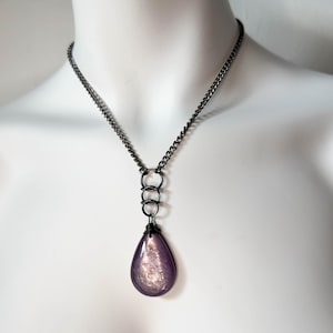 K&ouml;nnte beinhalten: Eine Halskette mit dunkler Metallkette und einem tropfenf&ouml;rmigen Anh&auml;nger. Der Anh&auml;nger hat eine perlmuttartige lila Farbe und ist an drei ineinandergreifenden Ringen aufgeh&auml;ngt. Die Halskette wird auf einer wei&szlig;en Schaufensterpuppe pr&auml;sentiert.