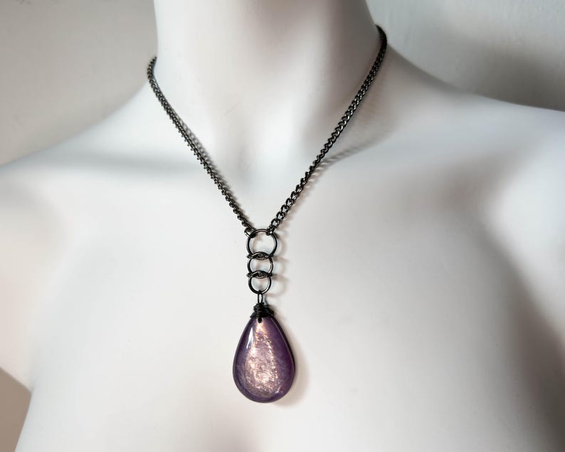 K&ouml;nnte beinhalten: Eine Halskette mit dunkler Metallkette und einem tropfenf&ouml;rmigen Anh&auml;nger. Der Anh&auml;nger hat eine perlmuttartige lila Farbe und ist an drei ineinandergreifenden Ringen aufgeh&auml;ngt. Die Halskette wird auf einer wei&szlig;en Schaufensterpuppe pr&auml;sentiert.
