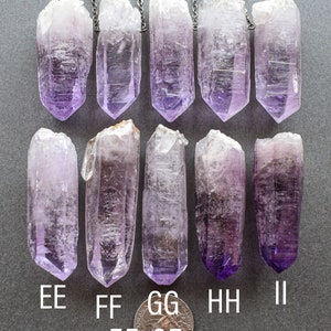 May include: Ten amethyst crystal points, each with a silver chain attached, are arranged in two rows of five. The crystals are approximately 55-65 mm in length. The crystals are a light purple color and have a clear, glassy appearance.