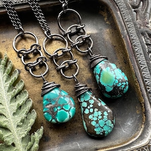 May include: Three teardrop-shaped turquoise pendants with dark matrix patterns, each wrapped in dark metal wire and suspended from dark metal chains featuring interconnected circular links, creating a cohesive set of necklaces.