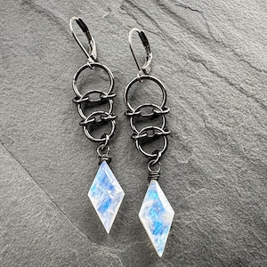 May include: A pair of dangle earrings featuring dark gunmetal-toned leverback hooks connected to three interlocking circular rings. Each earring terminates in a faceted, diamond-shaped moonstone pendant with iridescent blue and white flashes.