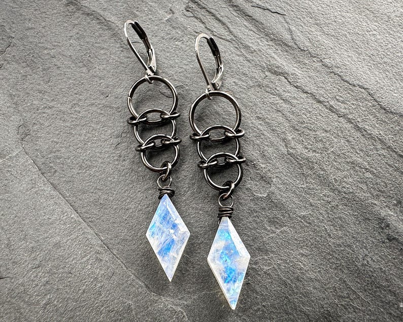 May include: A pair of dangle earrings featuring dark gunmetal-toned leverback hooks connected to three interlocking circular rings. Each earring terminates in a faceted, diamond-shaped moonstone pendant with iridescent blue and white flashes.