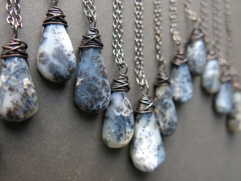 Puede incluir: Una colecci&oacute;n de colgantes de &oacute;palo dendr&iacute;tico en forma de l&aacute;grima, cada uno envuelto en alambre de cobre oscuro y suspendido de una cadena de metal a juego. Las piedras tienen una base blanca lechosa con inclusiones oscuras.