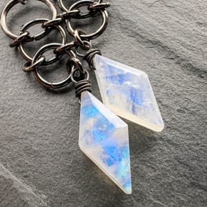 May include: Two diamond-shaped moonstone pendants suspended from dark gunmetal-colored interlocking jump rings. Each translucent gemstone displays a blue adularescence and is secured to the metal chain with wrapped wire detailing.