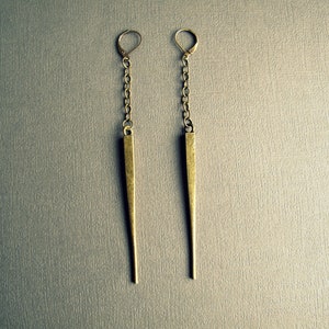 Pu&ograve; includere: Un paio di orecchini pendenti lunghi in metallo color ottone anticato. Ogni orecchino &egrave; composto da una chiusura a monachella collegata a una corta catena, che sostiene un pendente in metallo sottile, affusolato e a punta opaca.