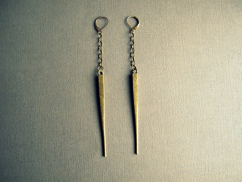 Pu&ograve; includere: Un paio di orecchini pendenti lunghi in metallo color ottone anticato. Ogni orecchino &egrave; composto da una chiusura a monachella collegata a una corta catena, che sostiene un pendente in metallo sottile, affusolato e a punta opaca.
