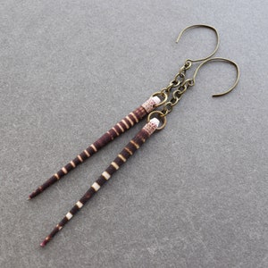 May include: A pair of earrings with brown and white striped, long, thin, teardrop-shaped pendants. The earrings are made with a bronze-colored metal hook.